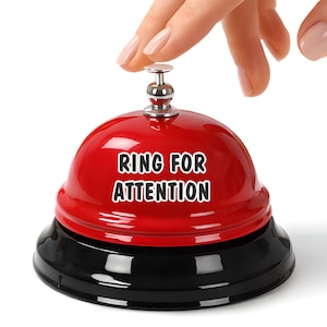 May include: A red service bell with a black base and a silver-colored top. The bell has the words "RING FOR ATTENTION" printed on the side. A hand is reaching for the top of the bell.