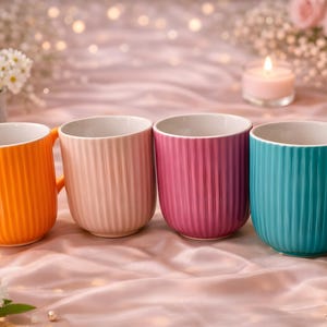 Può includere: Quattro tazze in ceramica in fila, ognuna di un colore diverso: arancione, rosa chiaro, magenta e turchese. Le tazze hanno una texture a coste e interni bianchi. Lo sfondo include fiori, una candela e un tessuto di raso rosa.