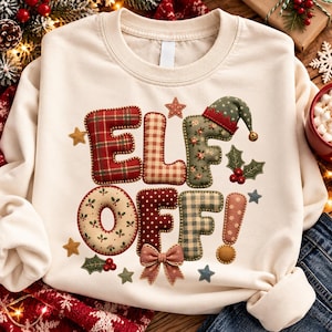 Op de afbeelding: Een crèmekleurige sweater met ronde hals en een opdruk met de tekst ELF OFF. De letters zijn ontworpen met diverse patronen, waaronder rood ruitjespatroon, beige gingham, groene bloemen en rode stippen, versierd met een strik.