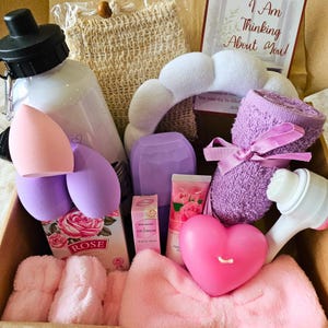 Op de afbeelding: Een geschenkdoos gevuld met zelfzorgartikelen. Inclusief een waterfles, make-upsponzen, een hoofdband, een gezichtsborstel, een kaars, lippenbalsem, een washandje en een luffa. De doos bevat ook een kaart met de tekst "I Am Thinking About You!"
