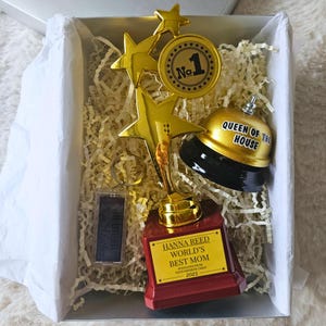 May include: A gift box containing a gold trophy, a bell, and a keychain. The trophy reads "HANNA REED WORLD'S BEST MOM" with the year 2025. The bell is labeled "QUEEN OF THE HOUSE". The box is filled with shredded paper.