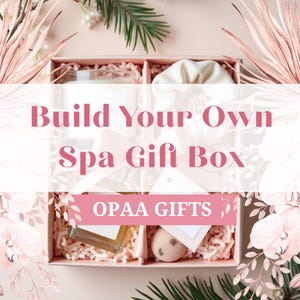 May include: A spa gift box with the text "Build Your Own Spa Gift Box" and "OPAA GIFTS". The box contains a candle, a small bag, a bottle, a lip balm, and a bath bomb, all arranged on pink paper filler.