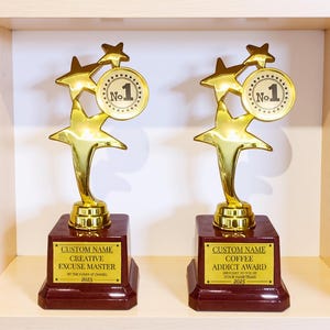 May include: Two gold-colored trophies with star-shaped accents and a "No. 1" medallion. The trophies sit on a dark red base with gold text that reads "Creative Excuse Master" and "Coffee Addict Award".