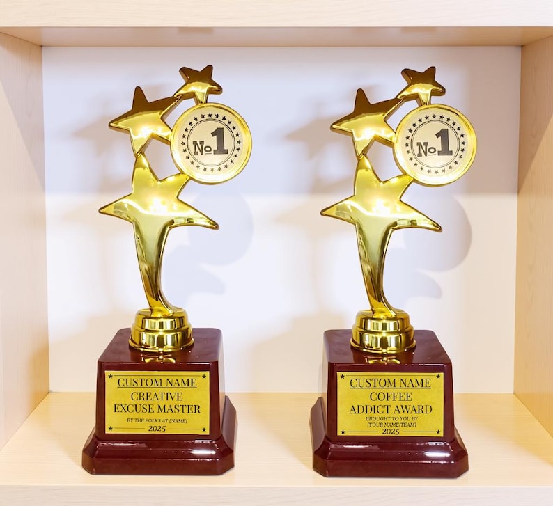 May include: Two gold-colored trophies with star-shaped accents and a "No. 1" medallion. The trophies sit on a dark red base with gold text that reads "Creative Excuse Master" and "Coffee Addict Award".