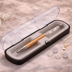 Op de afbeelding: Een goudkleurige pen met een zilveren punt en een transparante huls gevuld met iriserende kristallen, gepresenteerd in een zwarte etui met een transparant deksel. Een gouden ketting en diverse decoratieve stenen zijn ook zichtbaar.