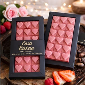 Può includere: Due barrette di cioccolato rosa a forma di cuore in scatole nere. Le barrette sono divise in quadrati, ognuno con un disegno a cuore. Le scatole sono etichettate "Casa Kakau Craft Chocolate". Sono visibili fragole e lamponi.