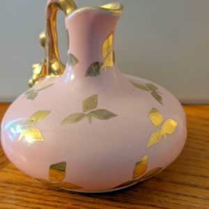 May include: A small, light pink ceramic pitcher featuring a gold-painted leaf pattern scattered across its rounded body. The pitcher has a gold-colored handle and a gold-trimmed rim, with a smooth, glossy finish throughout the entire piece.