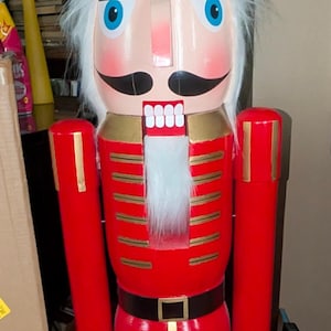 May include: A tall nutcracker figure wearing a black fuzzy bearskin hat with a gold chin strap. The figure has a red jacket with gold horizontal stripes, a black belt with a gold buckle, and black trousers. It features white hair, a mustache, and blue eyes.