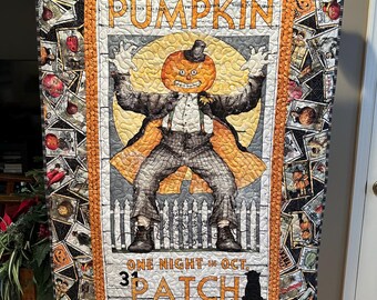 May include: A Halloween-themed quilt featuring a central image of a pumpkin-headed figure in a suit, arms raised. The quilt is bordered by vintage-style Halloween-themed postage stamps. Text reads "PUMPKIN PATCH" and "ONE NIGHT IN OCT. 31". A black cat is at the bottom.