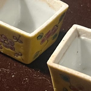 May include: Two square ceramic containers with a yellow exterior featuring hand-painted floral motifs in shades of pink and green. The interior of each vessel is white, and the rims show a slightly worn, off-white finish with visible texture.
