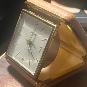 May include: A square Europa travel alarm clock with a gold-tone metal frame and a tan leather-like folding case. The white clock face features gold-tone hour markers, gold hands, and the text Europa and 2 Jewels printed on the dial.