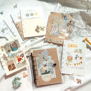 Puede incluir: Una colección de insertos de papel decorativos y un cuaderno con anillas. Los insertos presentan ilustraciones, incluyendo un gato, una mujer en un marco y arte con temática de café. El cuaderno es marrón con una cubierta transparente.