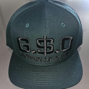May include: A dark teal baseball cap with a flat brim. The cap features the embroidered text "G$.O" in black, with the dollar sign incorporated into the design. Below the main text is the embroidered phrase "EVERYWHERE BUT NO WHERE".
