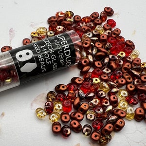 May include: A tube of Superduo Czech glass 2-hole seed beads in a mix of red, brown, and gold colors. The beads are scattered on a white surface.