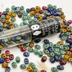 May include: A tube of colorful, two-hole glass seed beads. The beads are in shades of blue, green, red, and gold. The tube is labeled "Superduo Matubo Czech Glass 2-Hole Seed Beads".