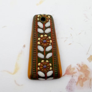May include: A rectangular pendant with a floral design. The pendant is primarily orange with a dark green border. The floral pattern includes white leaf shapes and brown flowers with purple centers. A small hole is at the top.