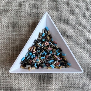 May include: A white triangular tray filled with a variety of small, colorful glass beads. The beads are in shades of black, gold, blue, and pink.
