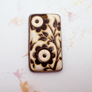 May include: Rectangular pendant with a floral design. The pendant features a cream-colored background with black floral patterns and a dark brown border. The design includes two large flowers and leafy vines. A small hole is at the top for stringing.