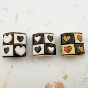 May include: Three ceramic beads with a checkerboard pattern of hearts. The beads are black, white, and yellow with red and orange hearts. The beads are arranged on a white surface with floral accents.