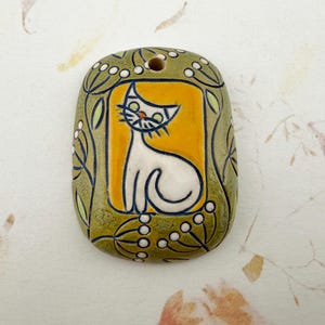 May include: A hand-painted ceramic pendant featuring a white cat on a yellow background. The cat is outlined in black, with green eyes and whiskers. The pendant is surrounded by a green border with floral and leaf details. The pendant has a hole at the top for hanging.