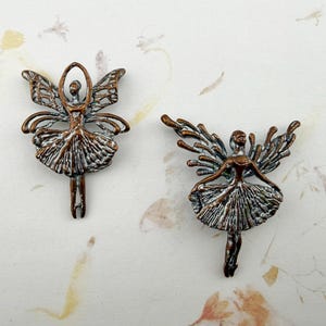 May include: Two bronze-colored fairy figurines. Each fairy has outstretched wings, a tutu, and arms raised above the head. The wings have an open, lacy design. The fairies are on a light-colored background.