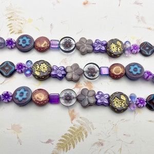 May include: Three strands of purple, brown, and clear glass beads with floral and geometric designs. The beads are strung on a clear thread.