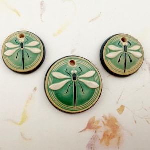 May include: Three round ceramic pendants featuring a raised white dragonfly design on a green background. Each pendant has a dark brown edge and a single hole at the top for hanging. The central pendant is larger than the two smaller ones.