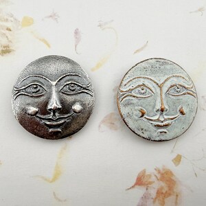 May include: Two round, smiling moon face embellishments. One is silver-toned metal, the other is painted with a light blue and bronze finish. Both have detailed facial features, including eyes, nose, and a smiling mouth. The background is a floral pattern.