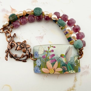 May include: A multi-strand bracelet featuring a rectangular focal piece with a hummingbird and floral print, accented by faceted pink and green beads, white pearls, and a copper-toned toggle clasp shaped like a flower with leaves.