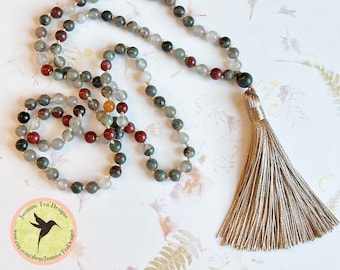 May include: A mala or prayer bead necklace made with green, grey, and red stones with a beige tassel. The necklace is laid out on a white surface with a floral pattern.