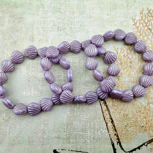 May include: Three circular bracelets composed of individual lavender-colored beads shaped like small seashells. Each bead features a series of raised, vertical ridges that mimic the texture of a scallop shell, creating a uniform, repeating pattern.