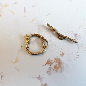 May include: Two gold-toned metal pieces shaped like branches. One piece is a ring and the other is a straight piece with a loop at one end.
