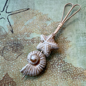 May include: A decorative skewer featuring a starfish and a nautilus shell, both in a cream and brown color scheme. The skewer has a looped handle made of thin, light-brown material. The background has a sea-themed design with starfish and coral.