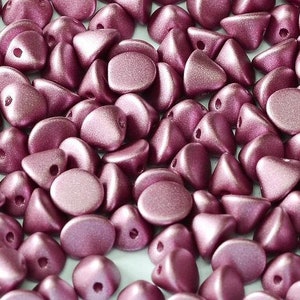 May include: A close-up of a pile of small, metallic pink beads. The beads are a mix of round and triangular shapes, and they have a matte finish.