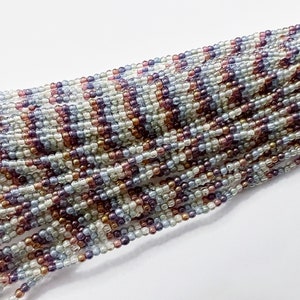 May include: A strand of seed beads in shades of purple, blue, and white. The beads are small and round, and they are arranged in a random pattern.
