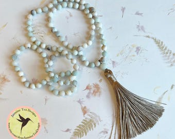 May include: A light blue and white beaded necklace with a long tassel. The necklace is made of small, round beads and has a long tassel at the end. The necklace is displayed on a floral background.