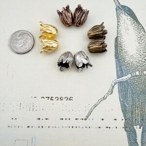 May include: A collection of eight tulip-shaped metal beads arranged in a circle, featuring metallic finishes in gold, silver, copper, and bronze tones. A US dime coin is positioned to the left for scale, showing the relative size of the beads.