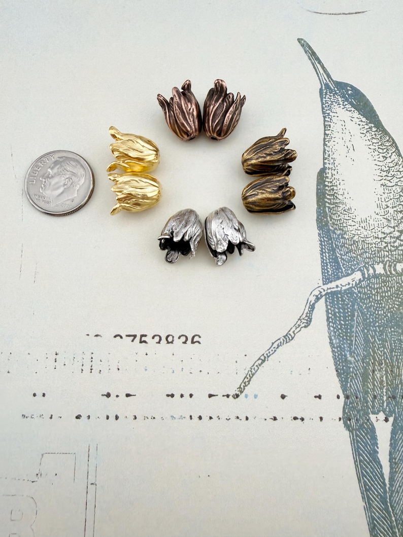 May include: A collection of eight tulip-shaped metal beads arranged in a circle, featuring metallic finishes in gold, silver, copper, and bronze tones. A US dime coin is positioned to the left for scale, showing the relative size of the beads.
