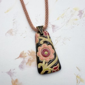 May include: A black ceramic pendant necklace with a floral design. The pendant features pink, red, and green flowers on a black background. The necklace is made with a copper chain.