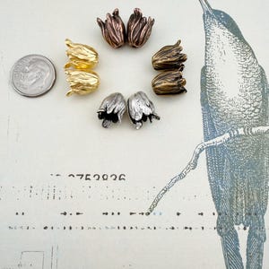 May include: Eight tulip-shaped metal beads arranged in a circle, featuring metallic finishes in gold, silver, copper, and bronze tones. A US dime coin is positioned to the left for scale, showing the relative size of these small floral components.