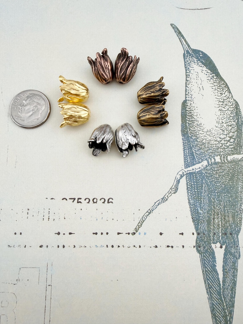 May include: Eight tulip-shaped metal beads arranged in a circle, featuring metallic finishes in gold, silver, copper, and bronze tones. A US dime coin is positioned to the left for scale, showing the relative size of these small floral components.