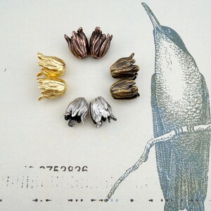 May include: A collection of eight metal tulip-shaped bead caps arranged in a circle. The set includes pairs in metallic finishes of bright gold, antique copper, dark bronze, and silver, each featuring detailed petal textures and a central hole.