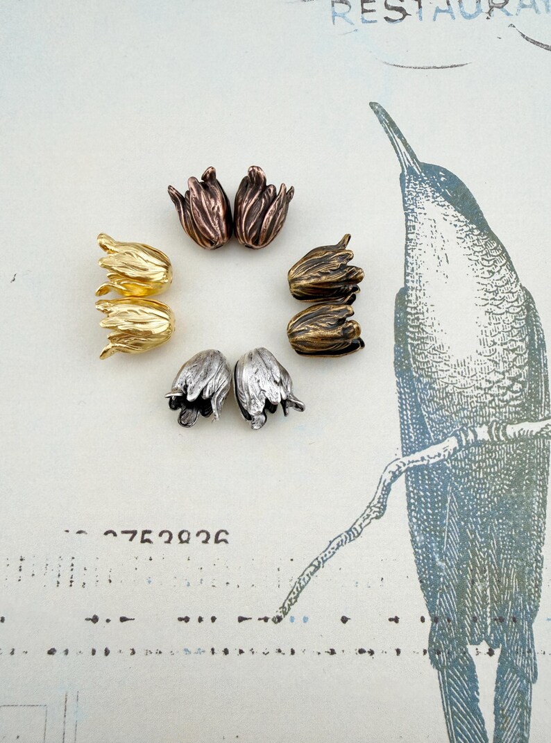 May include: A collection of eight metal tulip-shaped bead caps arranged in a circle. The set includes pairs in metallic finishes of bright gold, antique copper, dark bronze, and silver, each featuring detailed petal textures and a central hole.