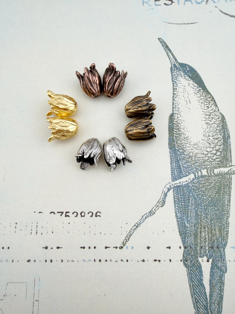 May include: A collection of eight metal bead caps shaped like tulip flowers, arranged in a circular pattern. The set includes pairs in metallic gold, copper, bronze, and silver tones, each featuring detailed petal textures and a hollow center.