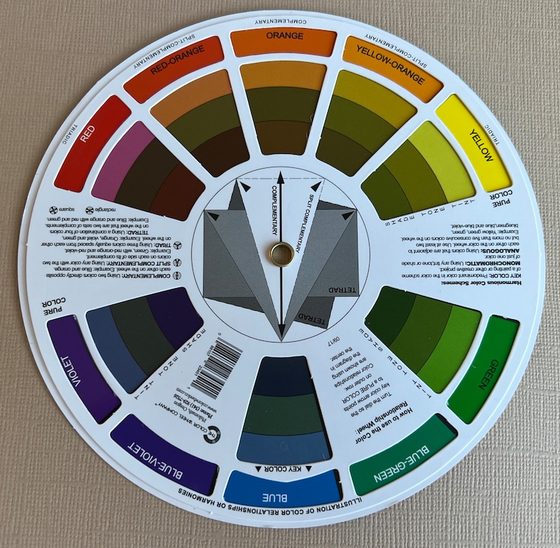 May include: A white circular color wheel with a gray center showing color relationships and harmonies. The wheel has 12 sections, each with a different color, including red, orange, yellow, green, blue, violet, and their corresponding shades and tints. The center of the wheel shows different color schemes, including complementary, split complementary, triadic, and tetradic.