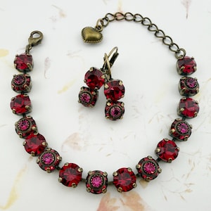May include: A vintage-style jewelry set featuring a bracelet and matching earrings. The set is made of antique brass-toned metal and adorned with vibrant red and pink crystals. The bracelet has a heart-shaped charm and adjustable chain.
