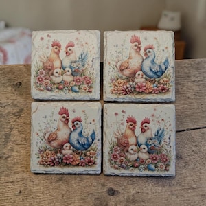 May include: Four square stone coasters featuring a watercolour design of chickens, chicks, and flowers. The chickens are in shades of peach, blue, and white, surrounded by pink and yellow flowers. The coasters are arranged on a wooden surface.