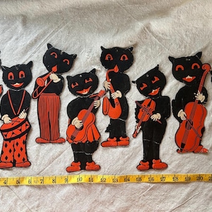 May include: A set of eight black and orange cat-shaped paper cutouts, each depicting a cat playing a different musical instrument, including an accordion, drum, trumpet, guitar, saxophone, violin, cello, and flute, arranged above a 23-inch ruler.