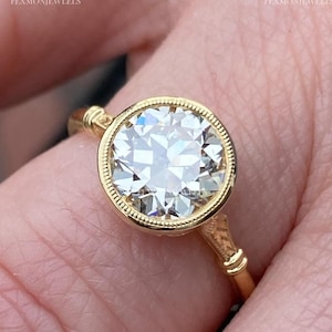 May include: A gold ring featuring a large, round, clear gemstone. The gemstone is set in a bezel setting with a detailed, textured edge. The ring's band has decorative elements near the gemstone and on the sides.
