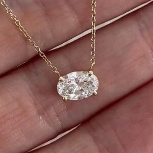 May include: A delicate gold necklace featuring an oval-cut diamond pendant. The diamond is set in a gold bezel and hangs from a fine gold chain. The necklace is displayed against a neutral background, highlighting the jewelry's sparkle.
